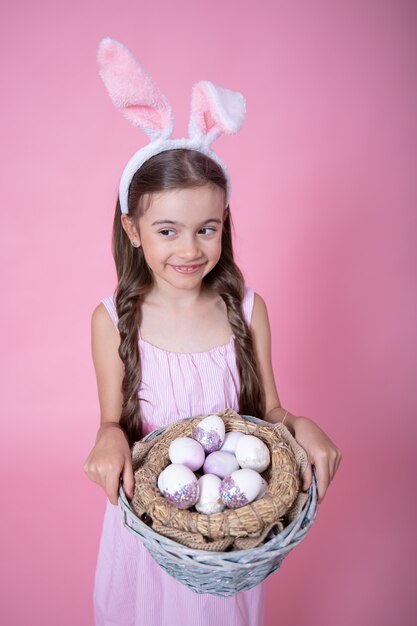 Petite fille avec des oreilles de lapin de Pâques posant tenant un panier avec des oeufs de Pâques festifs sur un mur rose se bouchent.