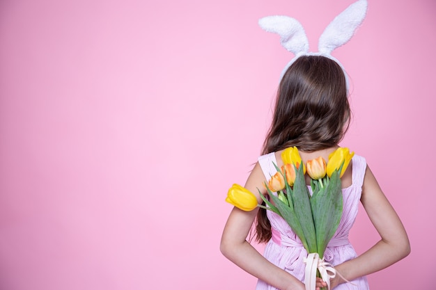 Une petite fille avec des oreilles de lapin de pâques tient un bouquet de tulipes dans ses mains derrière son dos sur un fond rose studio