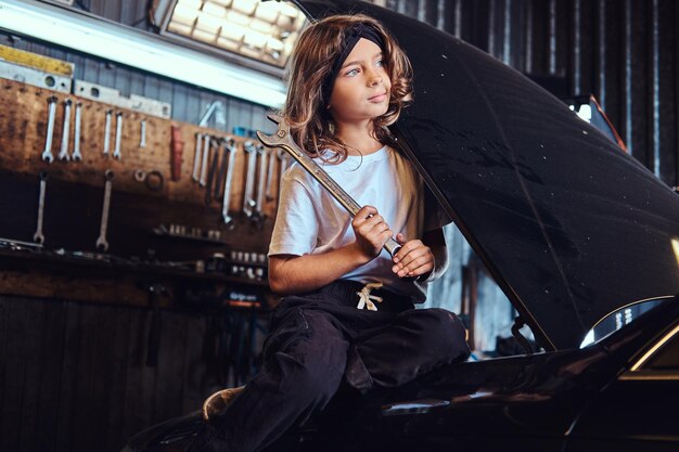 Une petite fille pensive est assise sur le capot de la voiture ouverte avec une grosse clé dans les mains.