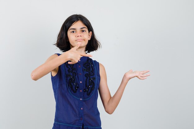 Petite fille pointant de côté, faisant semblant de tenir quelque chose en chemisier bleu et regardant le mécontentement.