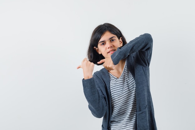 Petite fille pointant de côté avec les pouces en t-shirt, veste et à la confiance.