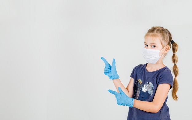 Petite fille pointant deux doigts regardant la caméra en t-shirt, gants et masque vue de face.