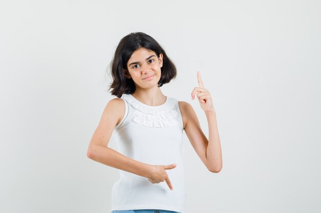 Petite fille pointant les doigts de haut en bas en chemisier blanc et à la joyeuse. vue de face.