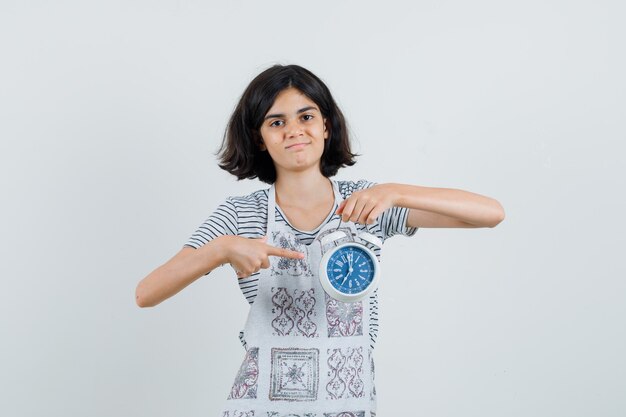 Petite fille pointant sur réveil en t-shirt, tablier et à la confiance,