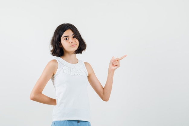 Petite fille pointant vers le côté en chemisier blanc, short et regardant heitant. vue de face.