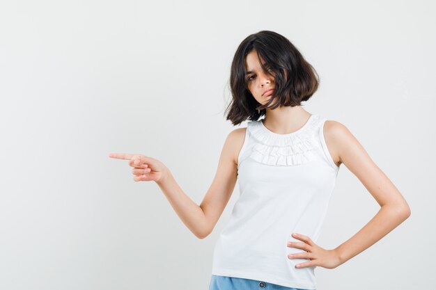 Petite fille pointant vers le côté en chemisier blanc, short et à la sérieuse. vue de face.