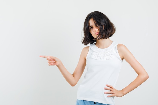 Petite fille pointant vers le côté en chemisier blanc, short et à la sérieuse. vue de face.