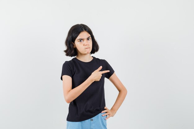 Petite fille pointant vers le côté en t-shirt noir, short et à la sérieuse, vue de face.