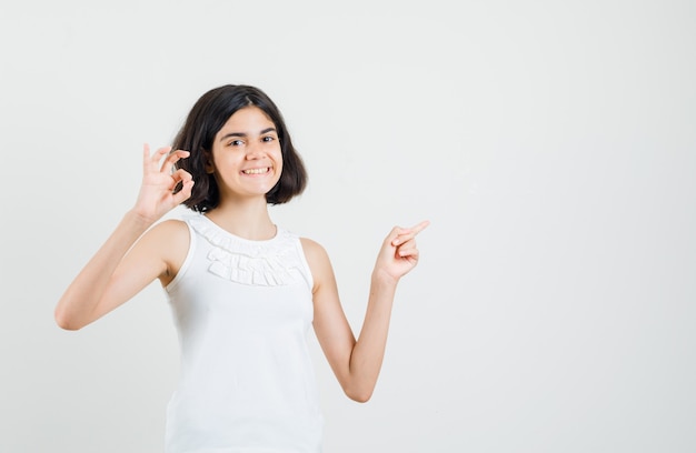 Petite fille pointant vers l'extérieur, montrant le signe ok en chemisier blanc et à la joyeuse. vue de face.