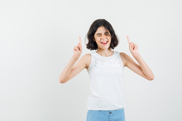 Petite fille pointant vers le haut en chemisier blanc, short et regardant heureux, vue de face.