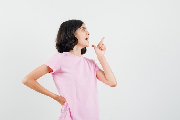 Petite fille pointant vers le haut en t-shirt rose et regardant heureux, vue de face.