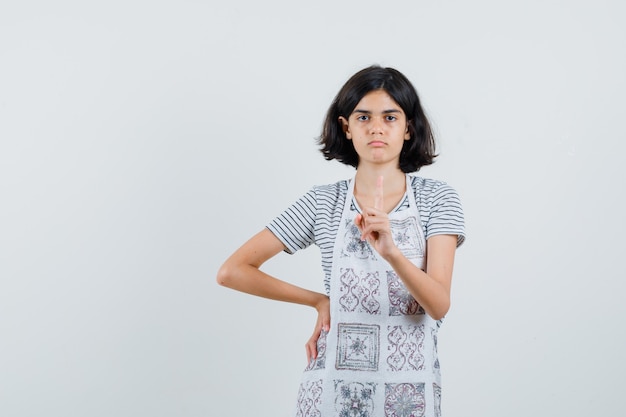 Petite fille pointant vers le haut en t-shirt, tablier et à la sérieuse