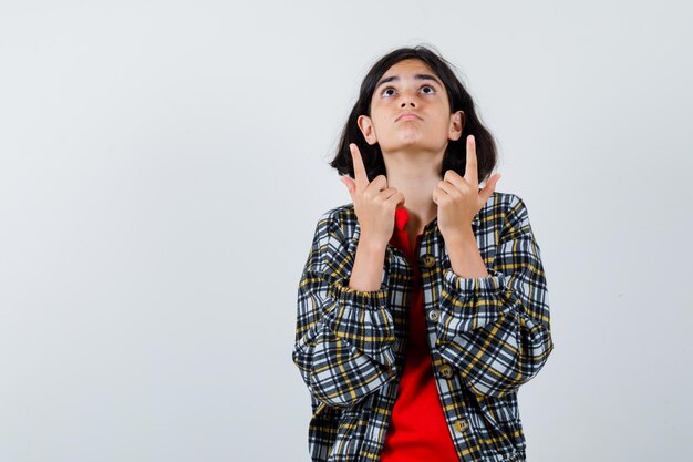 Petite fille pointant vers le haut tout en levant les yeux en chemise, veste et l'air concentré. vue de face. espace pour le texte