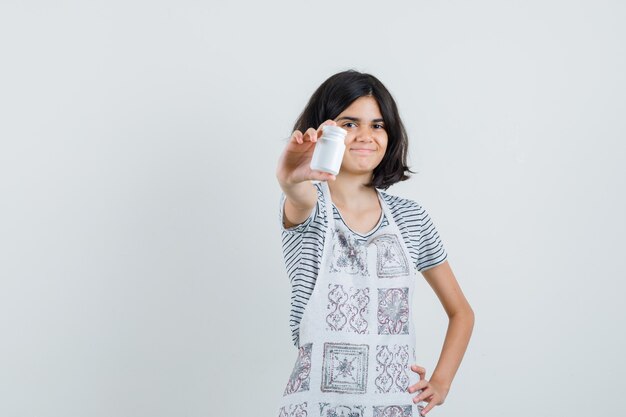 Petite fille présentant une bouteille de pilules en t-shirt, tablier et à la confiance.