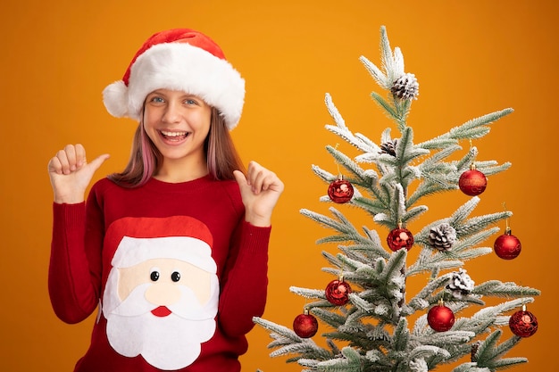 Photo gratuite petite fille en pull de noël et bonnet de noel heureux et surpris souriant joyeusement montrant les pouces vers le haut debout à côté d'un arbre de noël sur fond orange