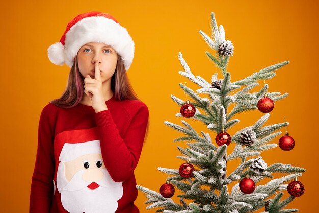 Petite fille en pull de noël et bonnet de noel regardant la caméra avec un visage sérieux faisant un geste de silence avec le doigt sur les lèvres debout à côté d'un arbre de noël sur fond orange