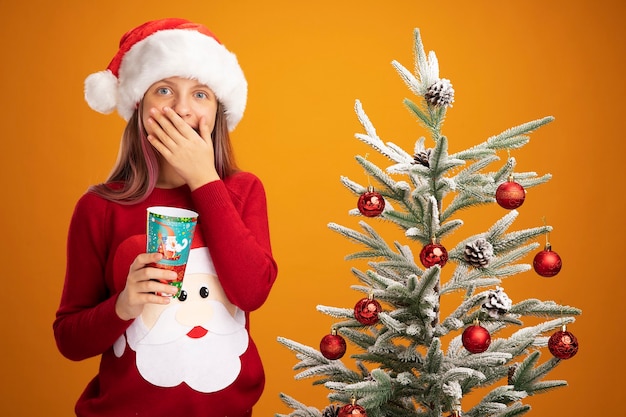 Petite fille en pull de noël et bonnet de noel tenant une tasse de papier coloré regardant la caméra heureuse et surprise couvrant la bouche avec la main debout à côté d'un arbre de noël sur fond orange