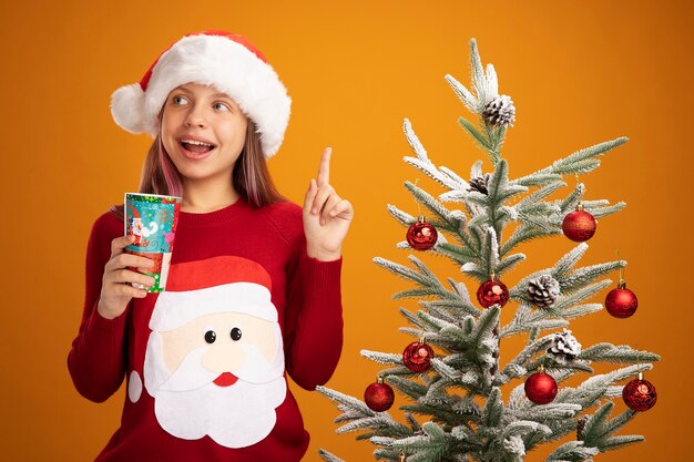 Petite fille en pull de Noël et bonnet de noel tenant une tasse de papier coloré