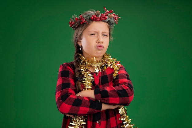 Petite Fille Rancunière Portant Une Couronne De Noël En Robe à Carreaux Avec Guirlandes Autour Du Cou Regardant La Caméra Avec Le Visage Froncé Avec Les Bras Croisés Debout Sur Fond Vert