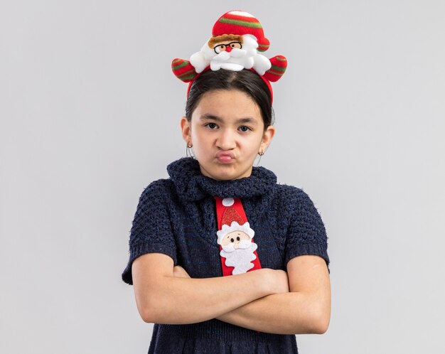 Petite fille rancunière en robe en tricot portant une cravate rouge avec une jante de Noël drôle sur la tête à la face en colère avec les bras croisés