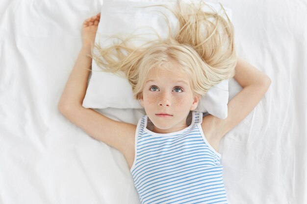 Petite fille réfléchie aux longs cheveux blonds levant les yeux, portant une chemise rayée, allongée sur un oreiller blanc, rêvant de nouveau vélo. Adorable fille reposante reposant sur un lit blanc dans la chambre des enfants