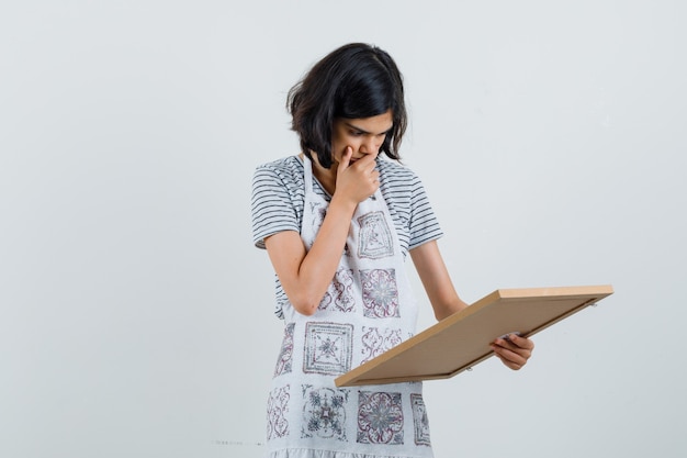 Petite Fille Regardant Un Cadre Vide En T-shirt, Tablier Et à La Surprise,