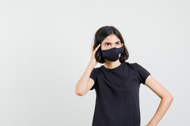 Petite fille regardant en t-shirt noir, masque et à la vue réfléchie, de face.