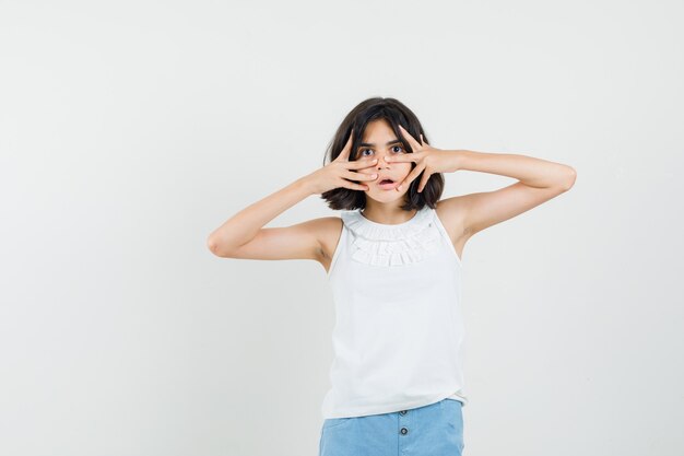 Petite fille regardant à travers les doigts en chemisier blanc, short et à la peur, vue de face.