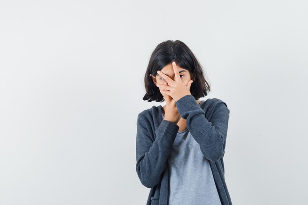 Petite fille regardant à travers les doigts en t-shirt, veste et à la curiosité,