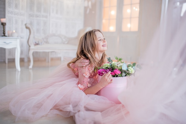 Petite fille en robe rose tient la boîte avec des roses assis sur le sol dans une chambre de luxe