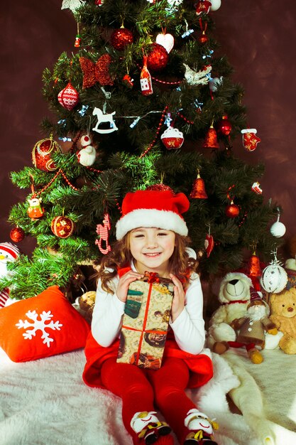 Petite fille en robe rouge est assis avec une boîte présente devant un arbre de Noël