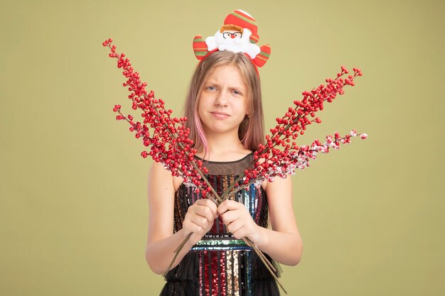 Petite fille en robe de soirée pailletée et bandeau avec le père Noël tenant des branches avec des baies rouges regardant la caméra faisant une bouche tordue avec une expression déçue debout sur fond vert