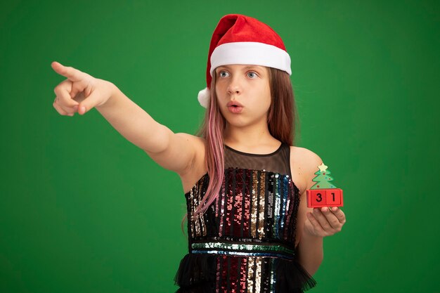 Petite fille en robe de soirée pailletée et bonnet de noel tenant des cubes de jouets avec la date du nouvel an regardant quelque chose de surpris pointant avec l'index sur le côté debout sur fond vert