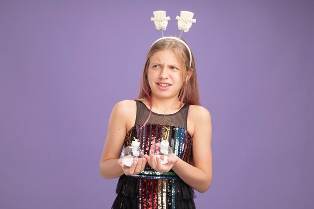 Petite fille en robe de soirée scintillante et bandeau drôle tenant des boules de Noël à côté confus et mécontent debout sur fond violet