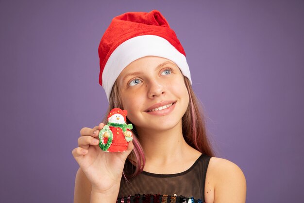Petite fille en robe de soirée scintillante et bonnet de noel montrant un jouet de noël levant avec le sourire sur le visage debout sur fond violet