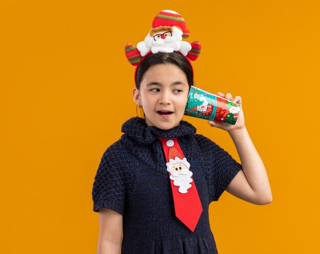 Petite fille en robe en tricot portant une cravate rouge avec jante drôle sur la tête tenant une tasse de papier coloré sur son oreille à l'intrigué