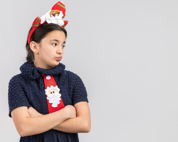 Petite fille en robe en tricot portant une cravate rouge avec jante de Noël drôle sur la tête à côté mécontent avec les bras croisés