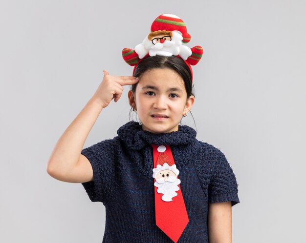 Petite fille en robe en tricot portant une cravate rouge avec jante de Noël drôle sur la tête à l'ennui faisant le geste du pistolet avec les doigts sur la tête