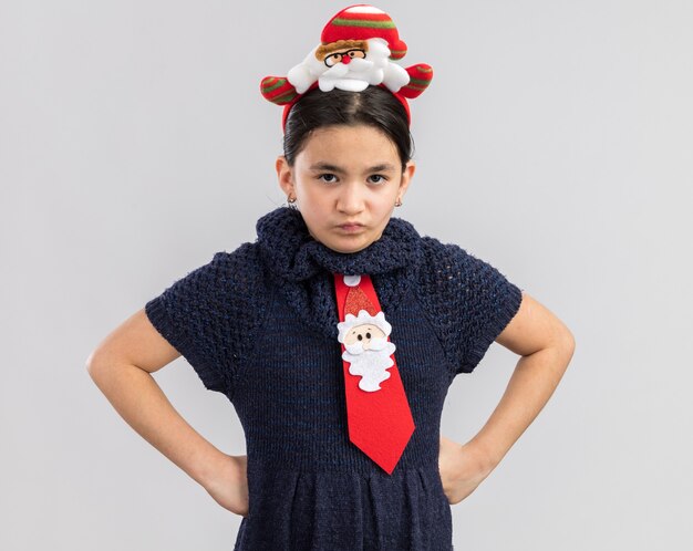 Petite fille en robe en tricot portant une cravate rouge avec jante de Noël drôle sur la tête à la face avec les sourcils froncés avec les bras à la hanche