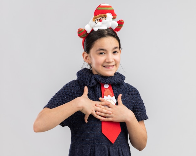 Petite fille en robe en tricot portant une cravate rouge avec une jante de Noël drôle sur la tête se tenant la main sur la poitrine se sentant reconnaissant