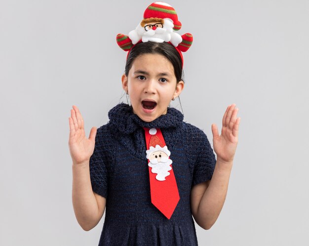 Petite fille en robe en tricot portant une cravate rouge avec jante de Noël drôle sur la tête à la surprise de faire un geste de grande taille avec les mains