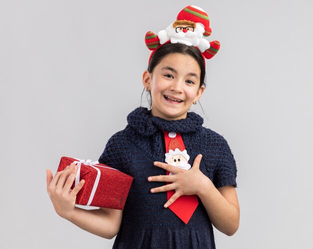 Petite fille en robe en tricot portant une cravate rouge avec jante de Noël drôle sur la tête tenant un cadeau de Noël à sourire joyeusement se sentir reconnaissant