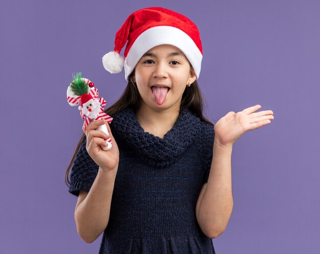 Petite fille en robe tricotée portant un bonnet de noel tenant une canne en bonbon de noël heureuse et joyeuse qui sort la langue debout sur un mur violet