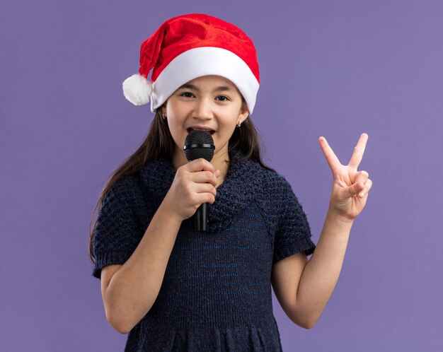 Petite fille en robe tricotée portant un bonnet de noel tenant un microphone chantant célébrant la fête de noël heureuse et positive montrant un signe v debout sur un mur violet