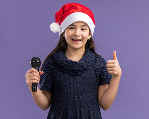 Petite fille en robe tricotée portant un bonnet de noel tenant un microphone heureux et positif montrant les pouces vers le haut debout sur un mur violet