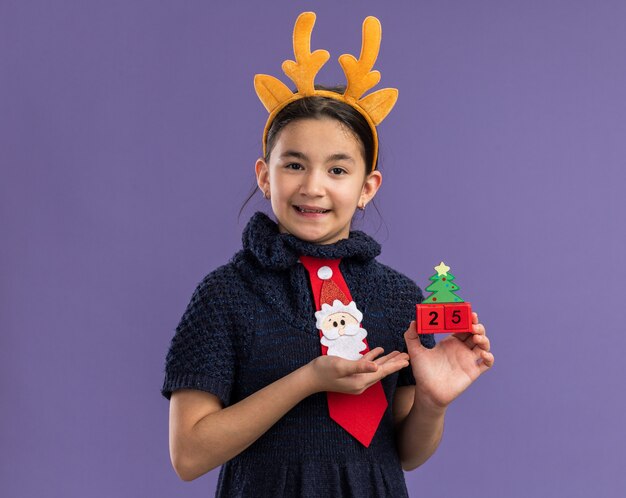 Petite fille en robe tricotée portant une cravate rouge avec un bord amusant avec des cornes de cerf sur la tête tenant des cubes de jouets avec la date de Noël présentant avec le bras un sourire heureux et positif debout sur un mur violet