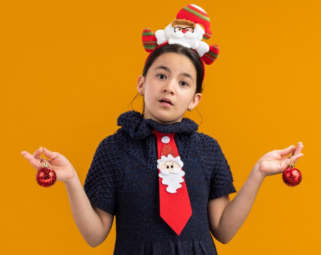 Petite fille en robe tricotée portant une cravate rouge avec un bord drôle sur la tête tenant des boules de noël confuses écartant les bras sur les côtés debout sur un mur orange