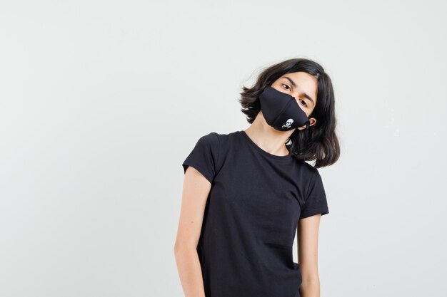 Petite fille s'inclinant la tête sur l'épaule en t-shirt noir, masque, vue de face.