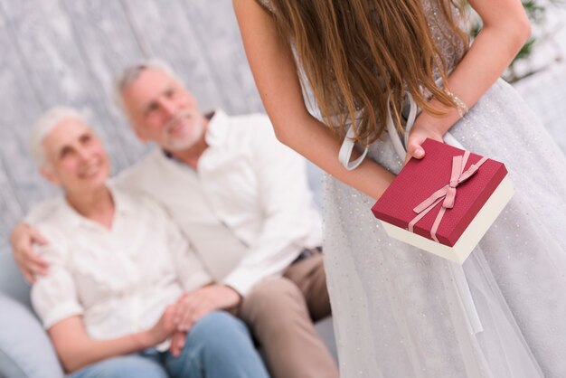 Petite fille se cachant un cadeau derrière son dos devant ses grands-parents assis sur un canapé