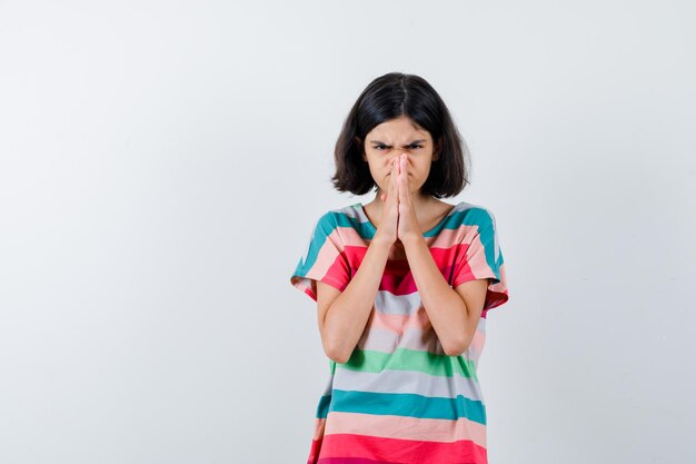 Petite fille serrant les mains en position de prière en t-shirt, jeans et l'air mécontent, vue de face.